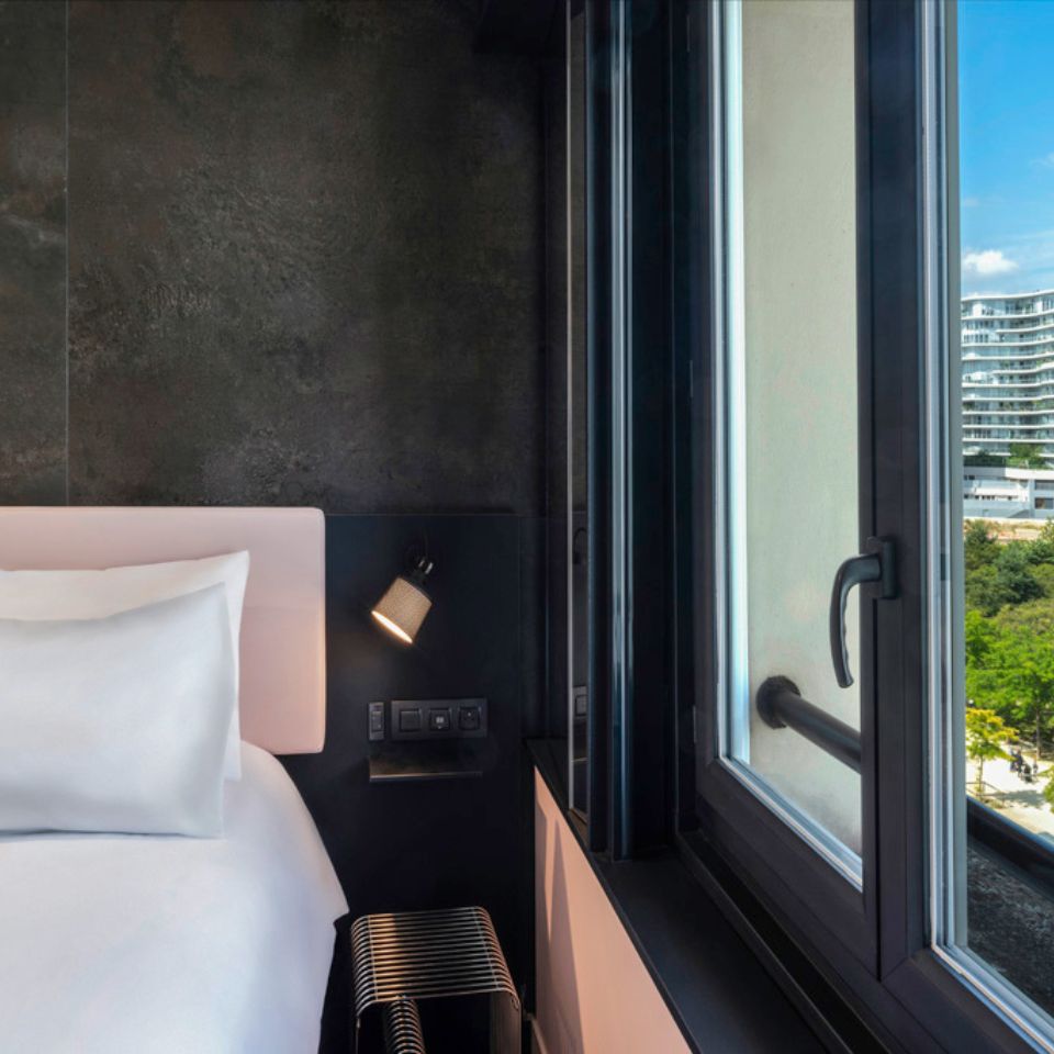 A hotel bed with a pink headboard against a black wall, the window shows a view of buildings and trees