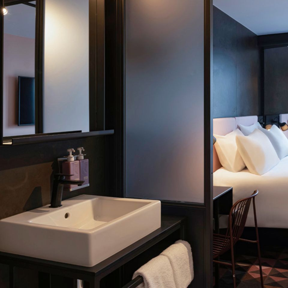 A washbasin with a mirror, in the background is a double bed and a small desk