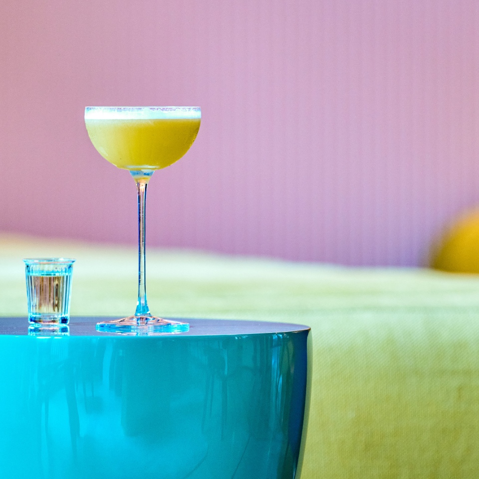 A vibrant yellow cocktail in a tall glass beside a small shot glass, resting on a glossy turquoise table with a colorful background.