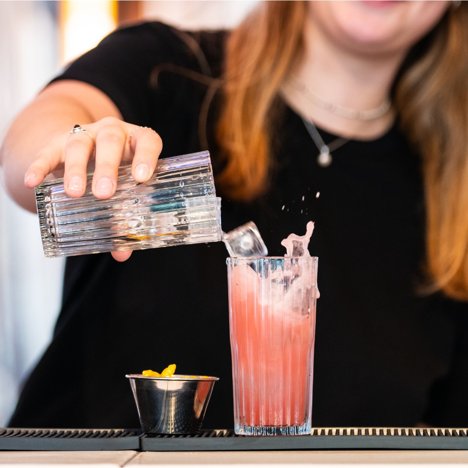 A bartender pours a clear liquid into a tall glass filled with a pink cocktail, with ice splashing, and a small bowl of orange garnish nearby.