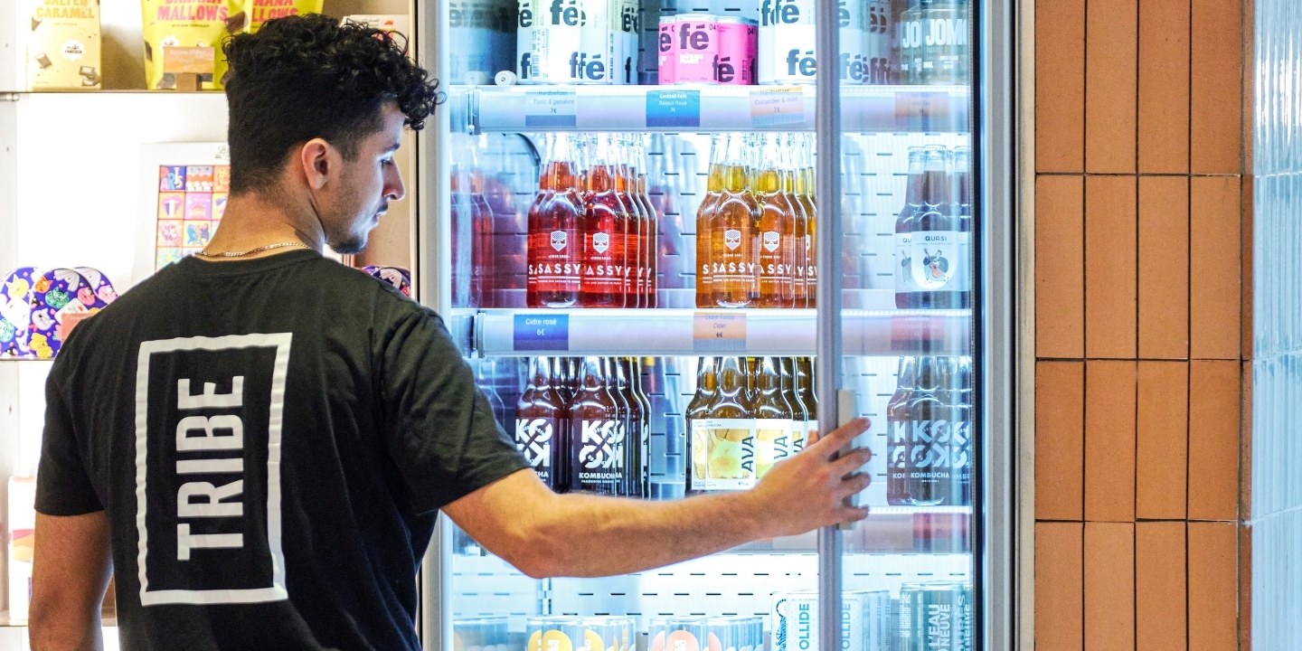 A person in a black "TRIBE" shirt is reaching into a refrigerated display filled with colorful beverage bottles.