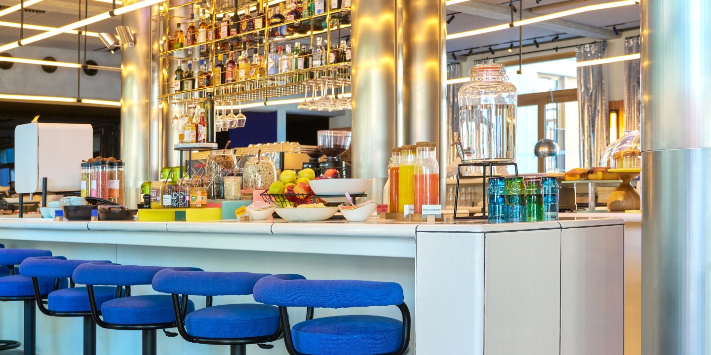 Modern bar with blue stools, a colorful assortment of drinks, fruits, and a well-stocked shelf of bottles under vibrant lighting.