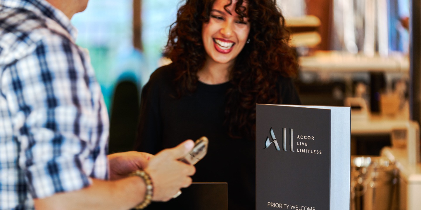 A person stands at a reception desk, engaging with a staff member. A sign for "Accor Live Limitless" and "Priority Welcome" is visible.