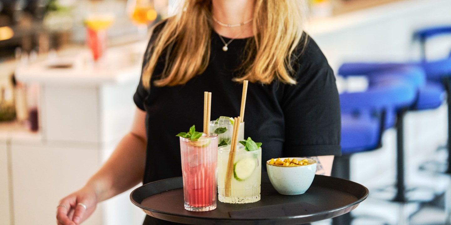 A server holds a tray with three colorful cocktails and a bowl of snacks, set against a vibrant bar backdrop.