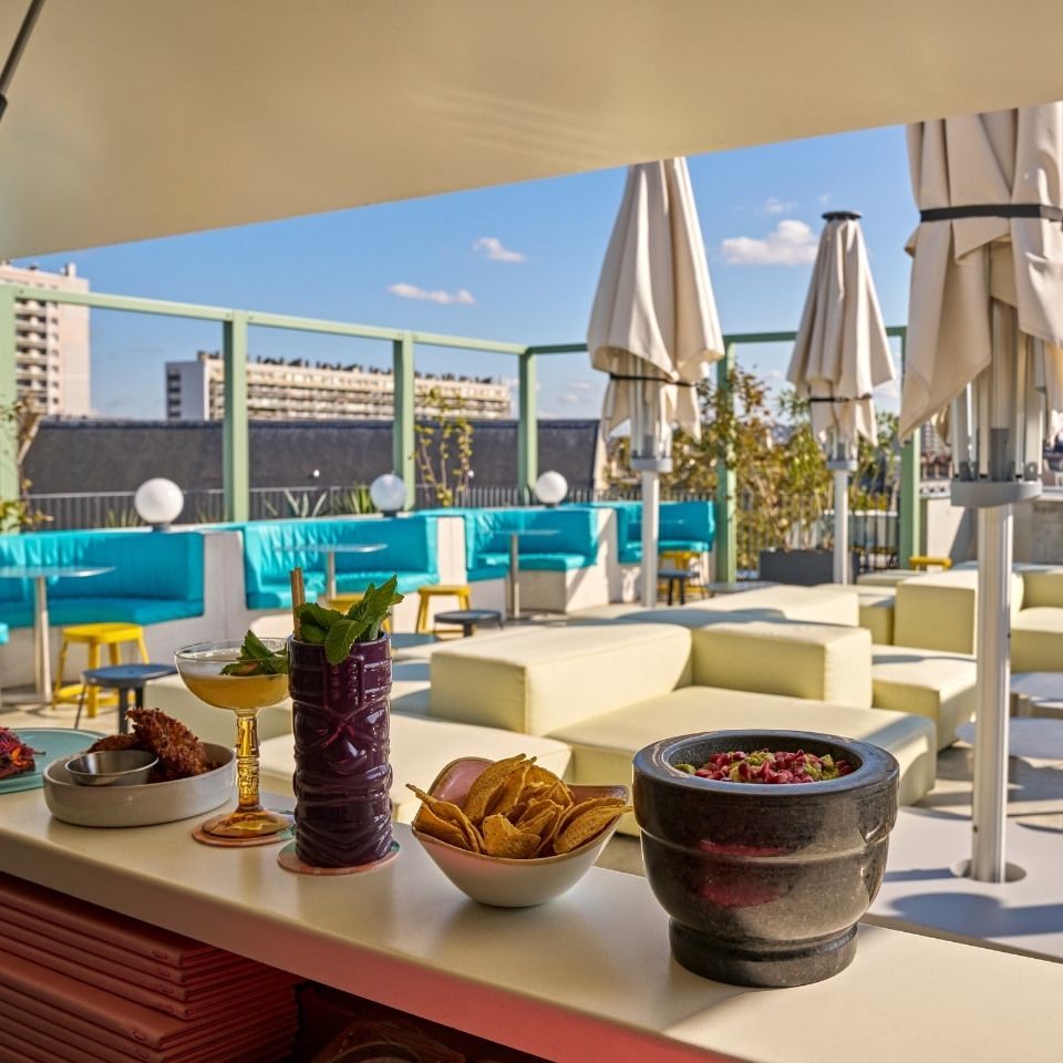 A vibrant rooftop bar scene featuring cocktails, snacks, and comfortable seating with city views under a clear blue sky.