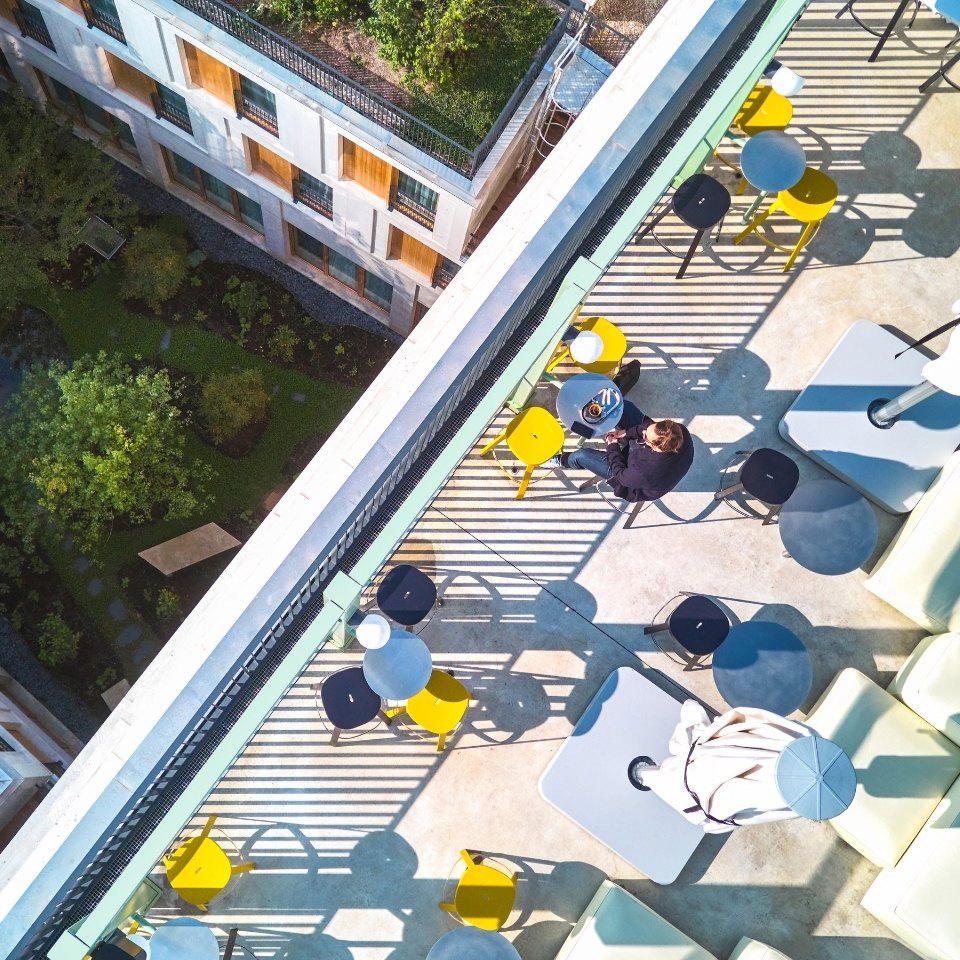 Aerial view of a rooftop terrace with colorful chairs and tables, featuring a person enjoying coffee amidst green surroundings.