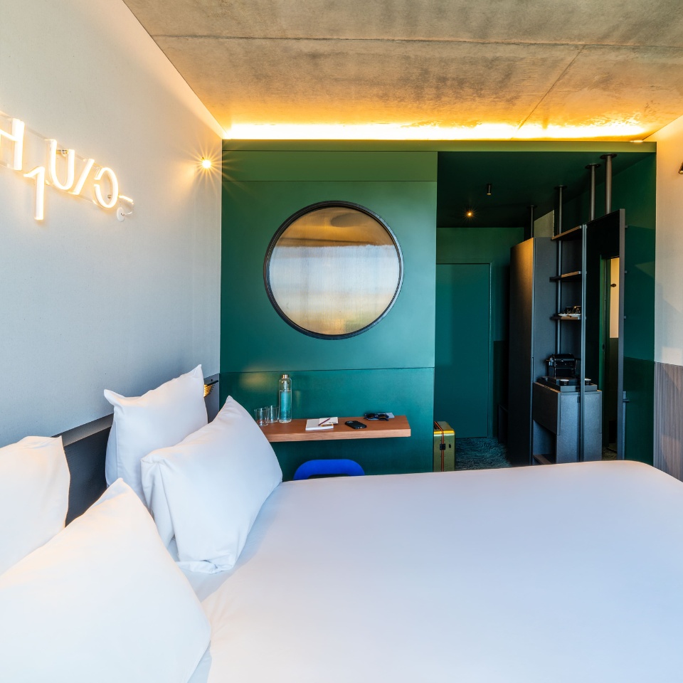 Cozy hotel room featuring a queen bed, green walls, circular window, modern decor, and a wooden desk with amenities.