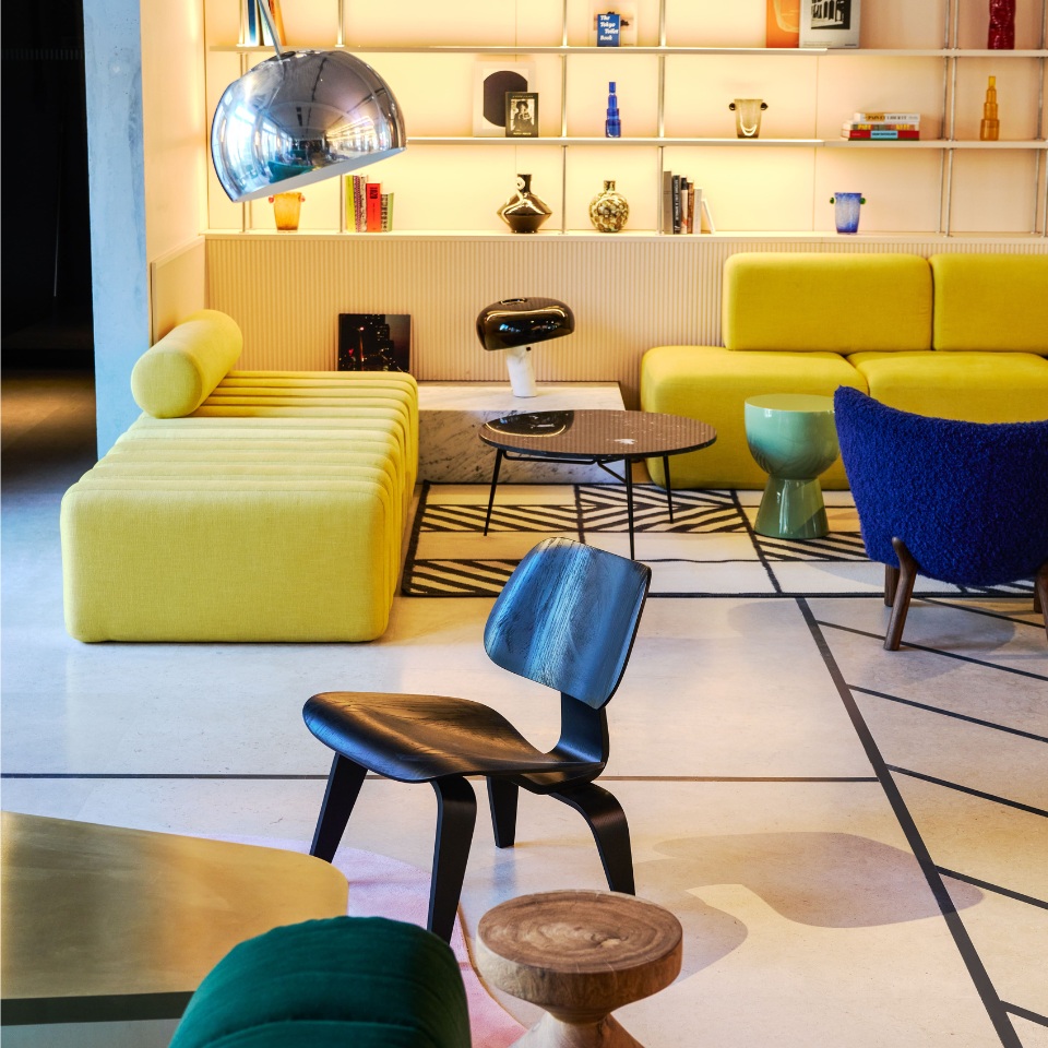 A colorful lounge with a yellow couch, blue and green chairs, modern decor, and a sleek metallic lamp, set against stylish shelving.