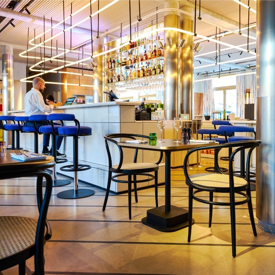 A modern restaurant interior with a sleek bar, stylish blue stools, and elegantly set tables, bathed in warm lighting.