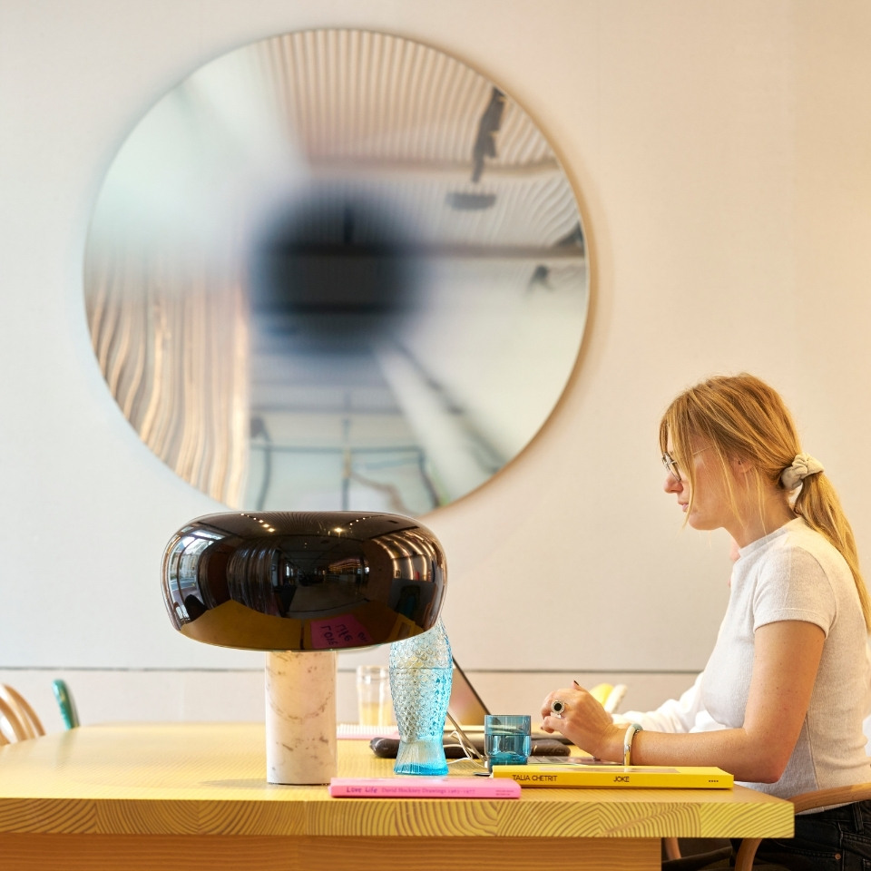 A woman writes at a wooden table adorned with a stylish lamp, colorful books, and a decorative glass, with a round mirror in the background.