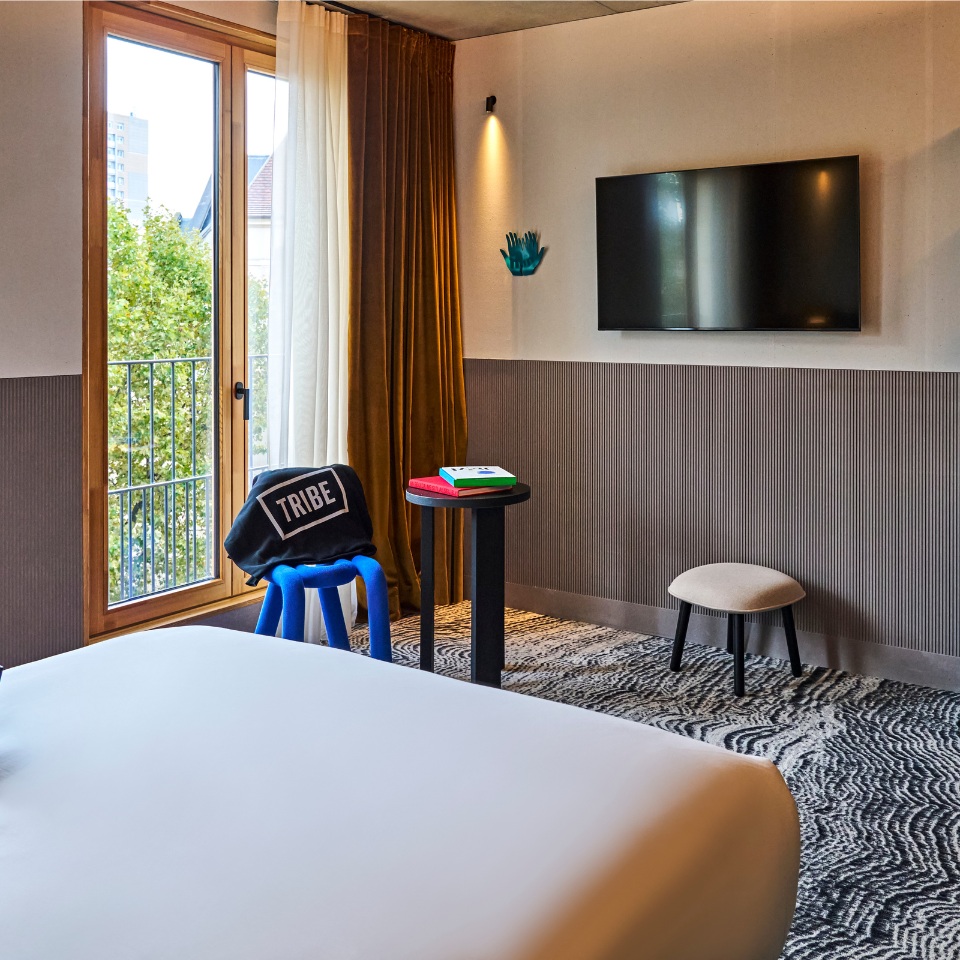 A hotel room featuring a bed, colorful furniture, a TV, and large windows with curtains, overlooking greenery outside.