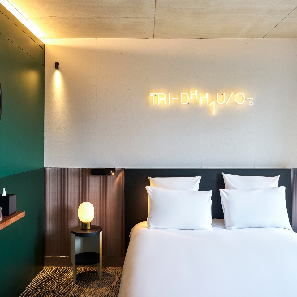 Modern hotel room featuring a double bed with white pillows, a stylish lamp, and neon signage on the wall. Dark green accent wall.