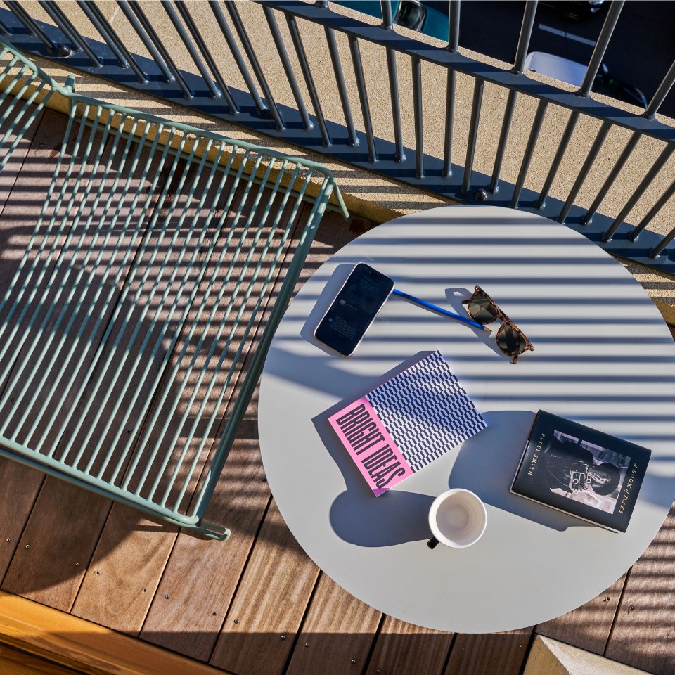 A sunlit balcony scene featuring a circular table with a coffee cup, books, sunglasses, and a smartphone.