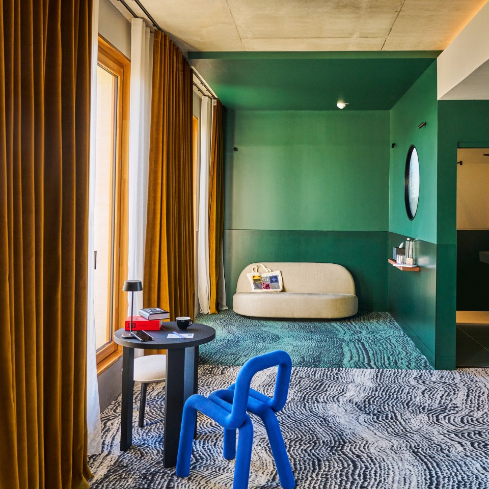 A stylish interior with green walls, mustard curtains, a blue chair, a small black table, and a cozy beige sofa by a bright window.