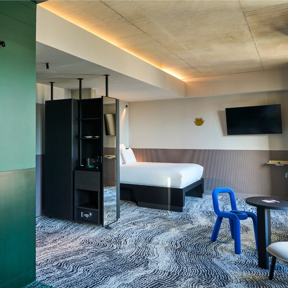 Stylish hotel room featuring a comfortable bed, modern decor, and a blue chair, with warm lighting and a television.