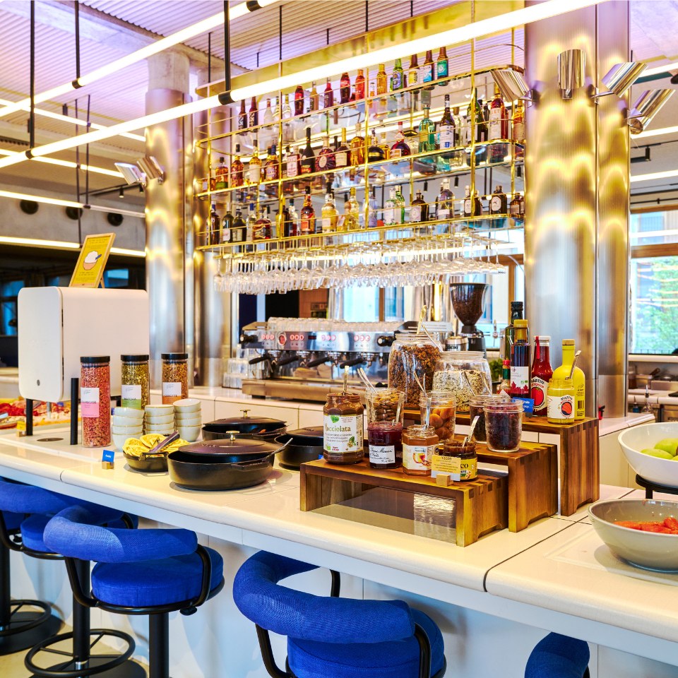 A breakfast bar with colorful bottles, jars of condiments, and snacks on a sleek countertop, accompanied by blue bar stools and bright lighting.