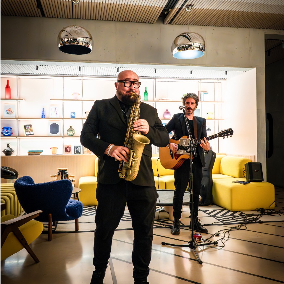 Two musicians perform in a stylish setting; one plays a saxophone while the other strums an acoustic guitar, surrounded by modern decor.