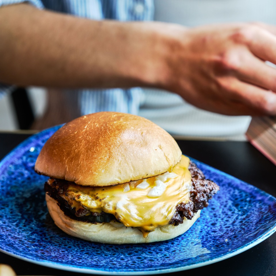 A close-up of a juicy cheeseburger with melted cheddar cheese, served on a blue plate, with a hand reaching for it in the background.
