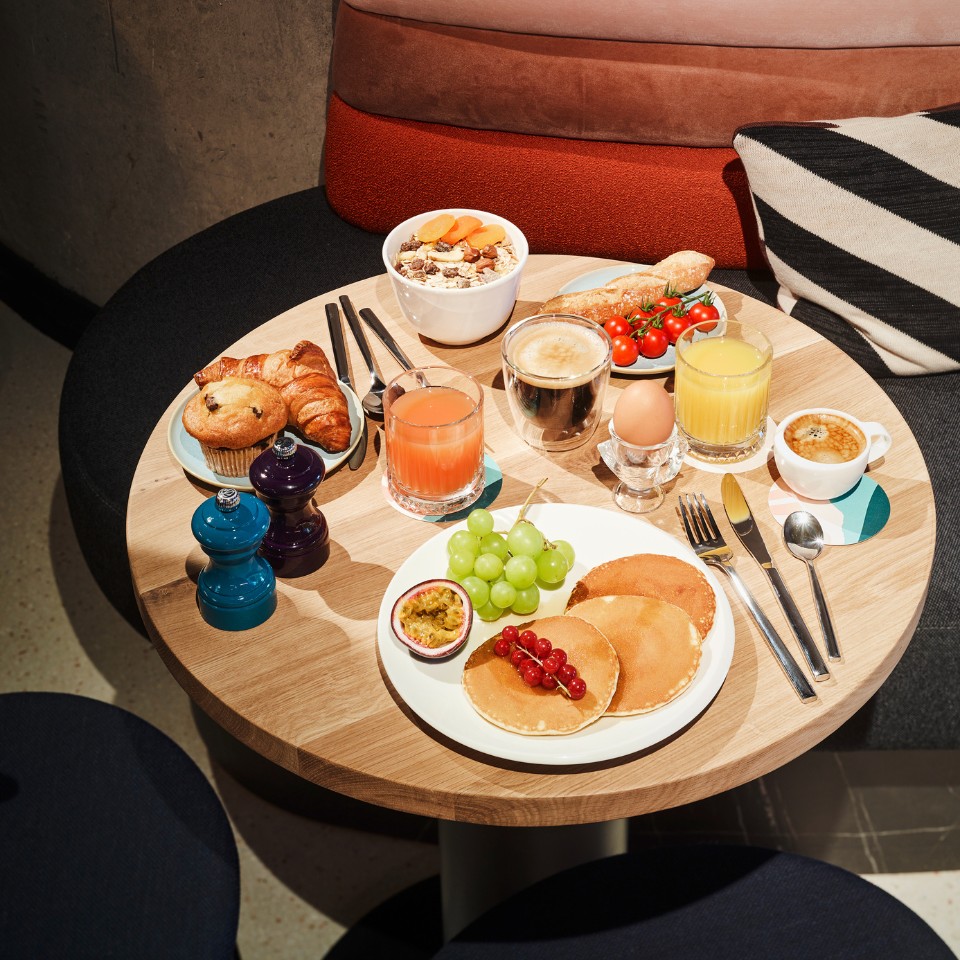 A wooden, round table with several dishes on it, like pancakes, croissants, an egg and coffee and juices