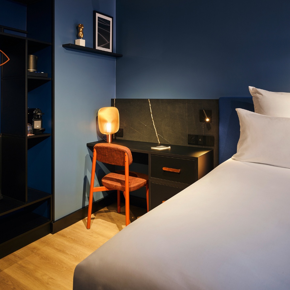 Small black desk next to a double bed, with an orange chair in front of it. The wall is blue