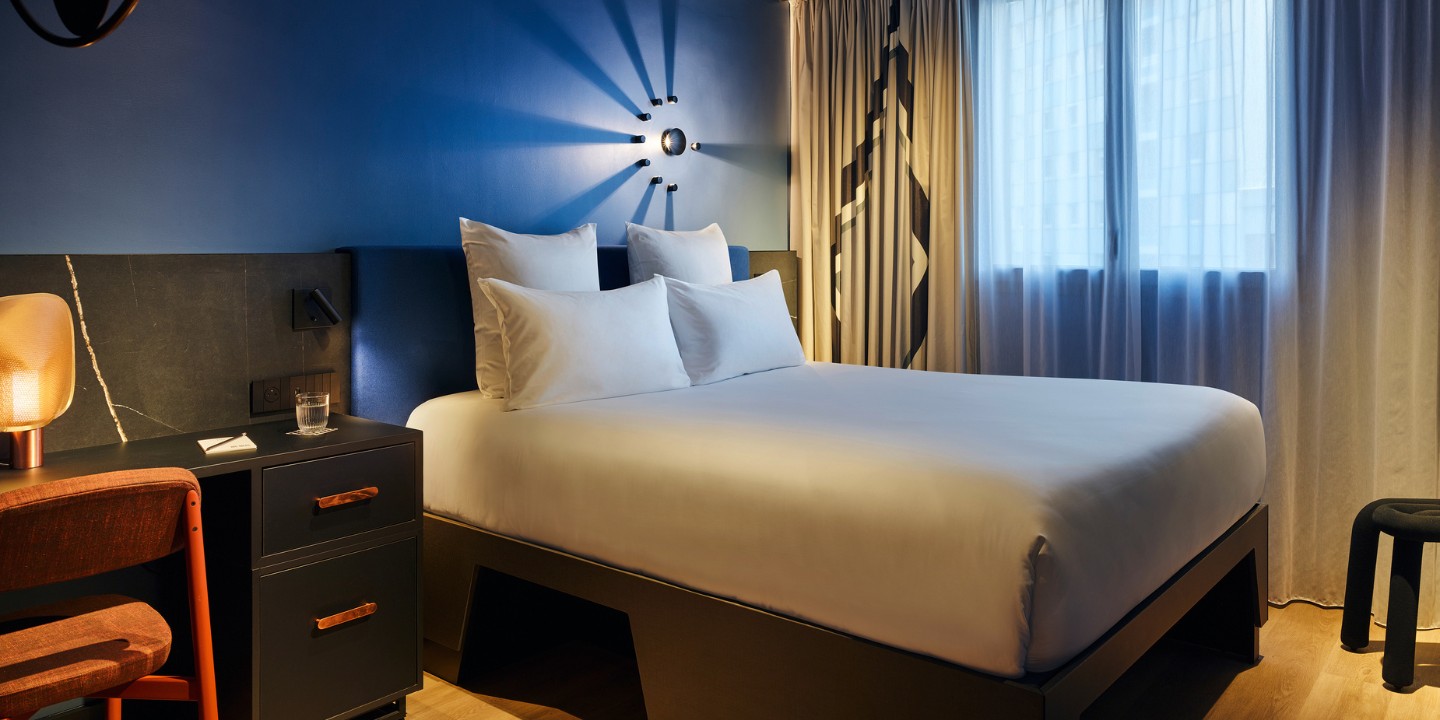 Double bed with white sheets and a blue headboard against a blue wall, to the left is a small black desk