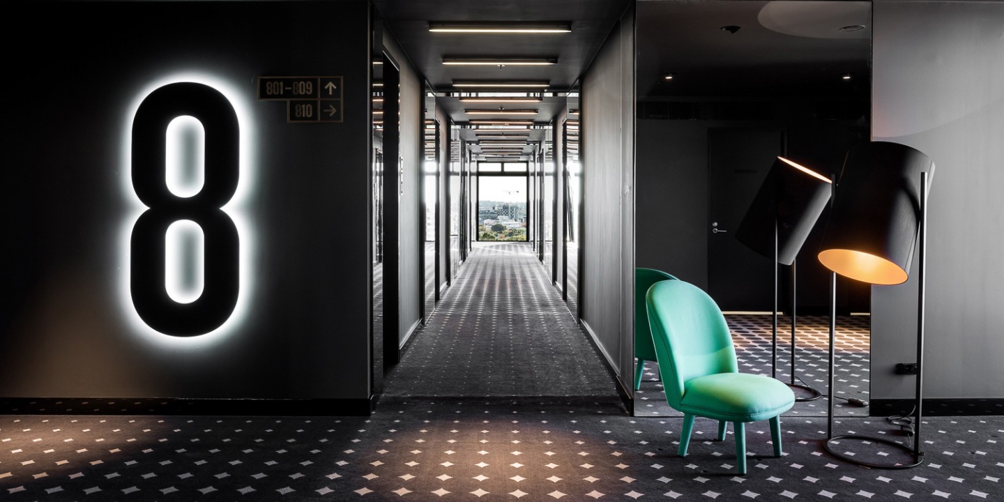 Hallway in the hotel with dark walls and carpet, on the wall is a large 8 and on the right is a green chair with a lamp.