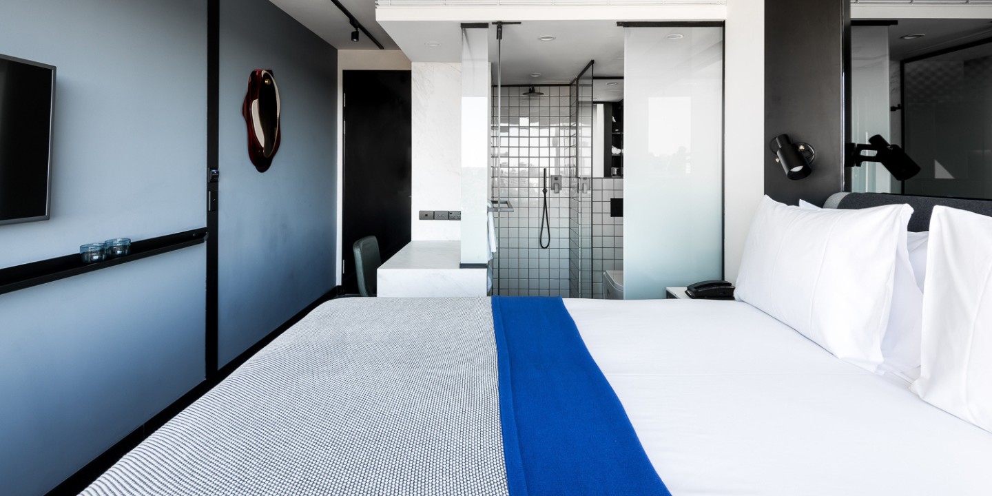 double bed with white linen in a hotelroom with blue walls, in the background the bathroom is visible