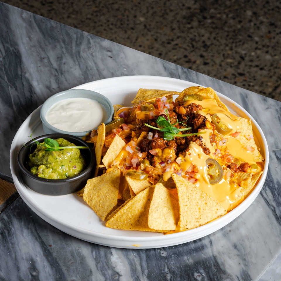 Loaded nachos with cheese, minced meat and jalapenos on a plate. There are two dips, guacamole and sour cream, next to it.