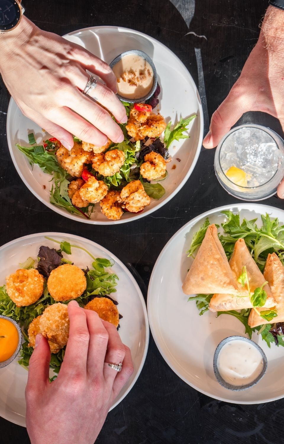 Three plates of appetizers—including breaded bites and pastry triangles—are placed on a dark table with dipping sauces. Hands are reaching for the food.