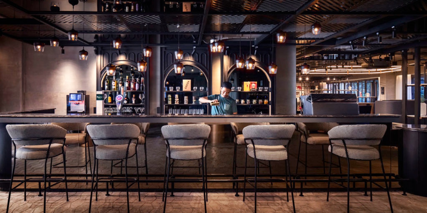 the TRIBE Express bar with a barkeeper serving a drink behind it and bar chairs in front of it. there are several liquers in the background