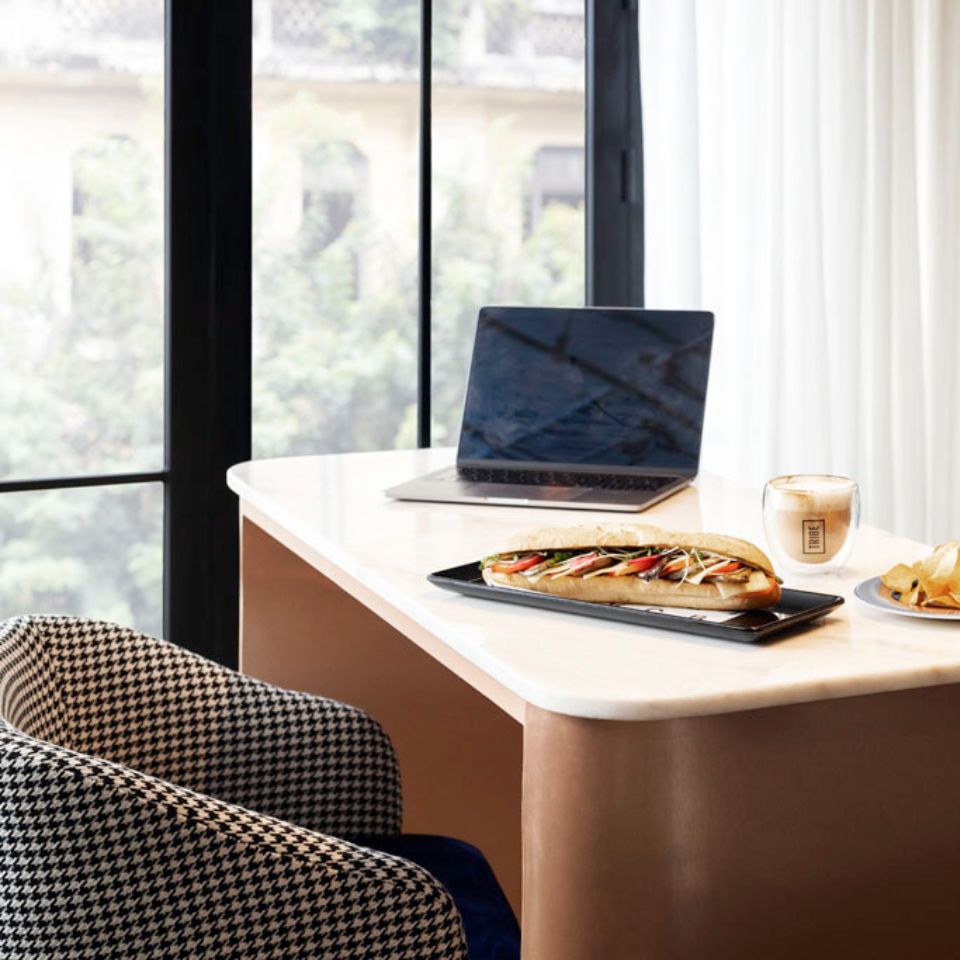 A workspace with an armchair, a table with a laptop on it. There is a baguette and some coffe next to the laptop