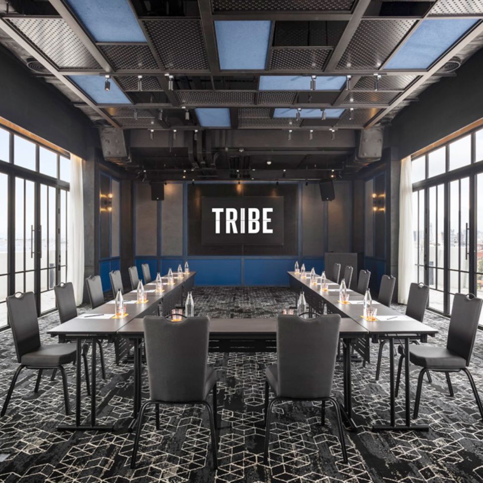 Several chairs and tables set up classroom style in a room with a dark floor, floor-to-ceiling windoes and a blue wall, on which there is a large screen