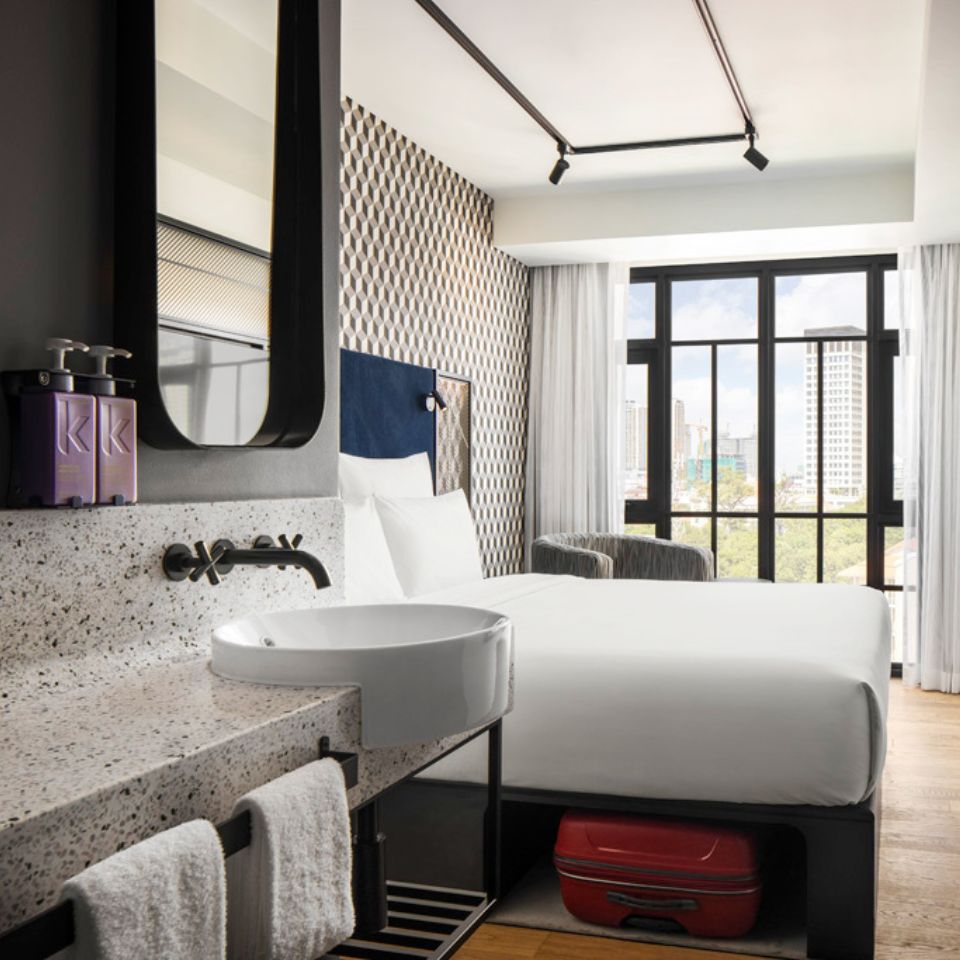 Guest room with a double bed, a blue headboard, an armchair and a washstand. The floor to ceiling window shows the city of Phnom Penh