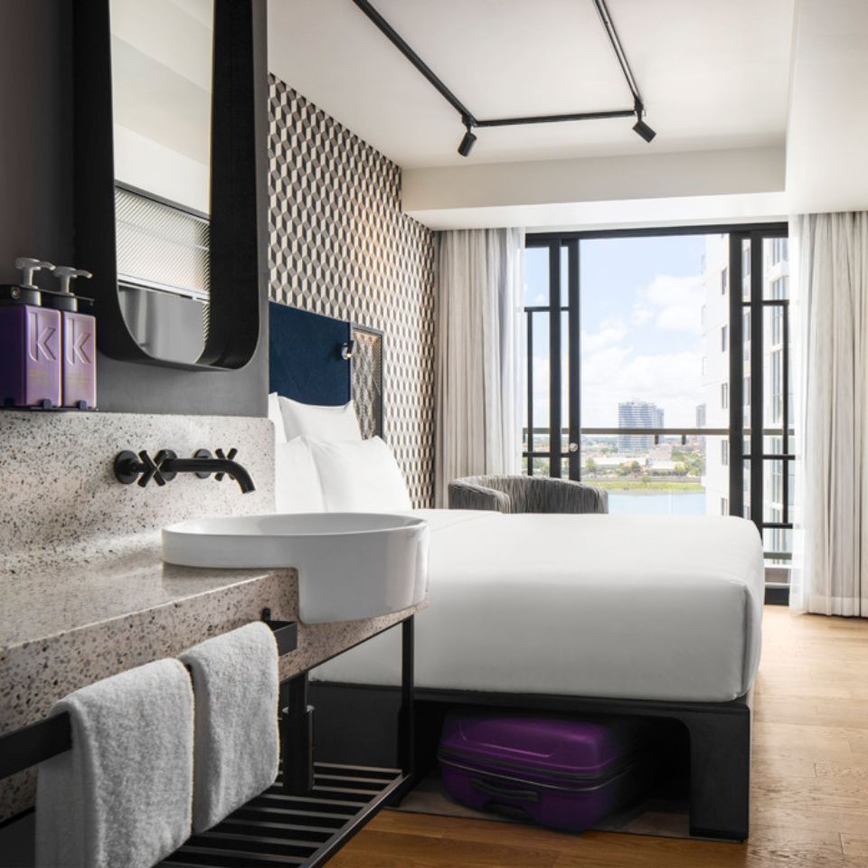 Guest room with a double bed, a blue headboard, an armchair and a washstand. The floor to ceiling window shows the city of Phnom Penh