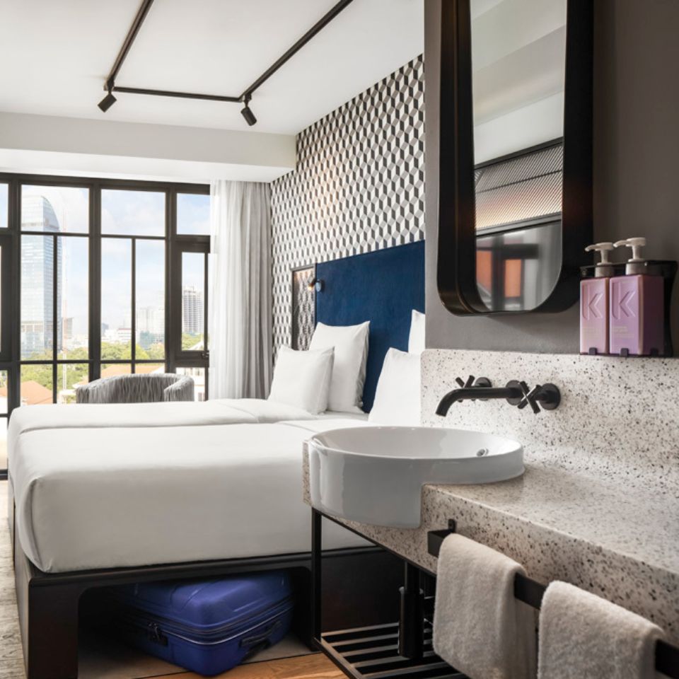 Guest room with a double bed, a blue headboard, an armchair and a washstand. The floor to ceiling window shows the city of Phnom Penh