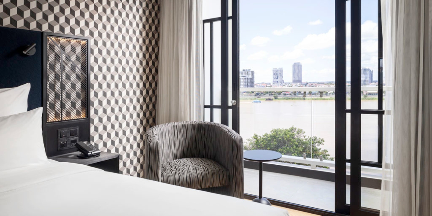 Guest Room with a bed with white linen, a gra armchair, a small black table and a floor-to-ceiling door which lead to a balcony with a view of the Mekong river and Phnom Penh city