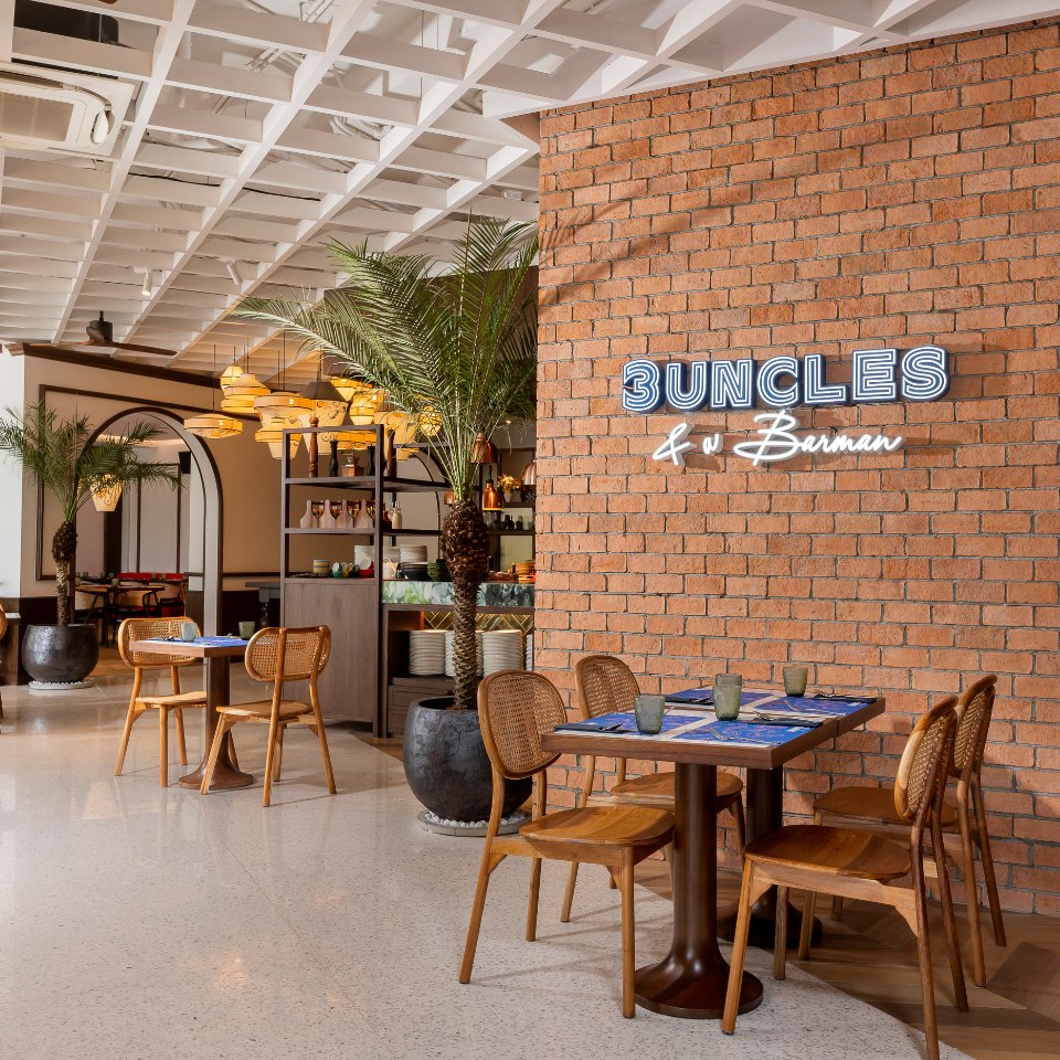 Modern café interior with exposed brick wall featuring neon "3 Uncles & a Barman" sign. Cozy wooden tables, wicker chairs, and potted palms add warmth.