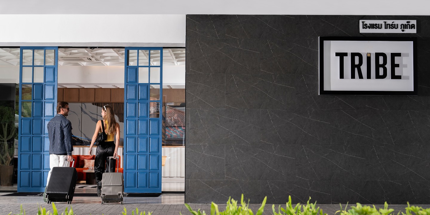 A couple stands at a stylish entrance, pulling luggage, framed by vibrant blue doors and sleek modern architecture.