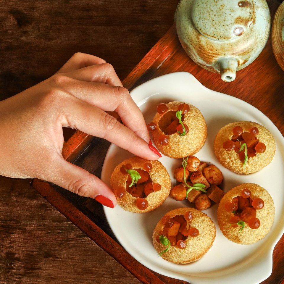 A hand with red nails reaches for a crispy, round snack on a white plate. The snacks are filled with garnishes and a side of small, diced fillings. A ceramic teapot sits nearby, creating a cozy and inviting scene.
