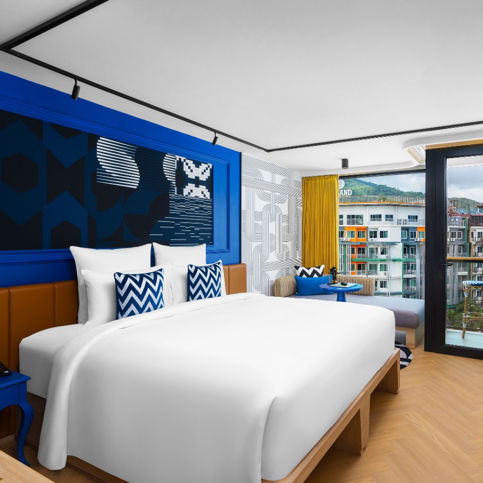 Modern hotel room featuring a king bed, vibrant blue accents, geometric patterns, and a balcony with colorful city views.