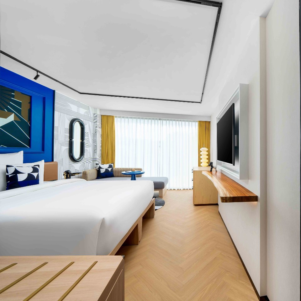 A modern hotel room featuring a large bed, stylish decor, a comfortable seating area, and bright natural light from floor-to-ceiling windows.