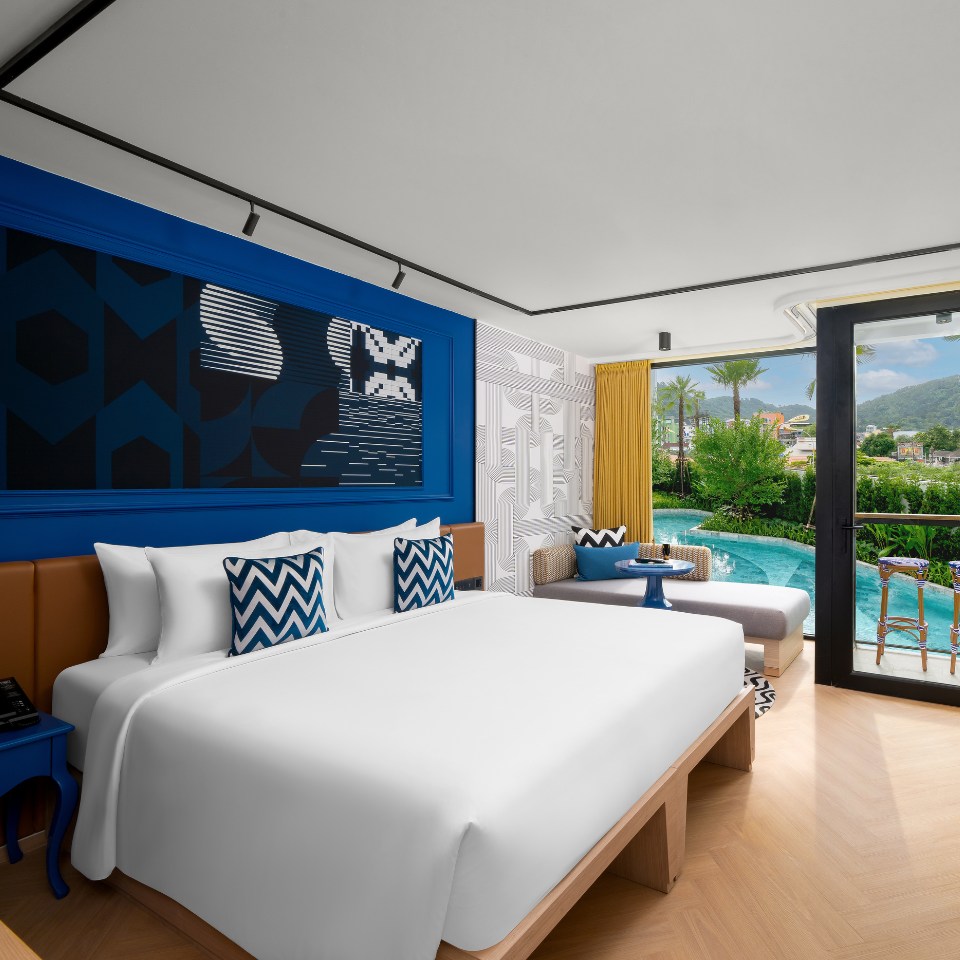 Modern hotel room featuring a white bed with blue accents, vibrant wall art, and a view of a pool and greenery through large windows.