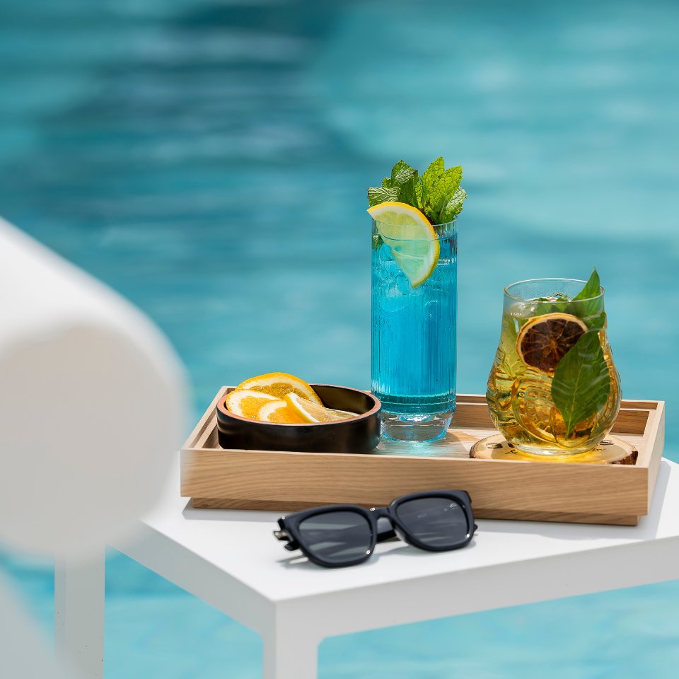 Two colorful drinks, one blue with lemon and mint, the other amber with citrus and basil, sit on a tray by a pool, with sunglasses beside them.