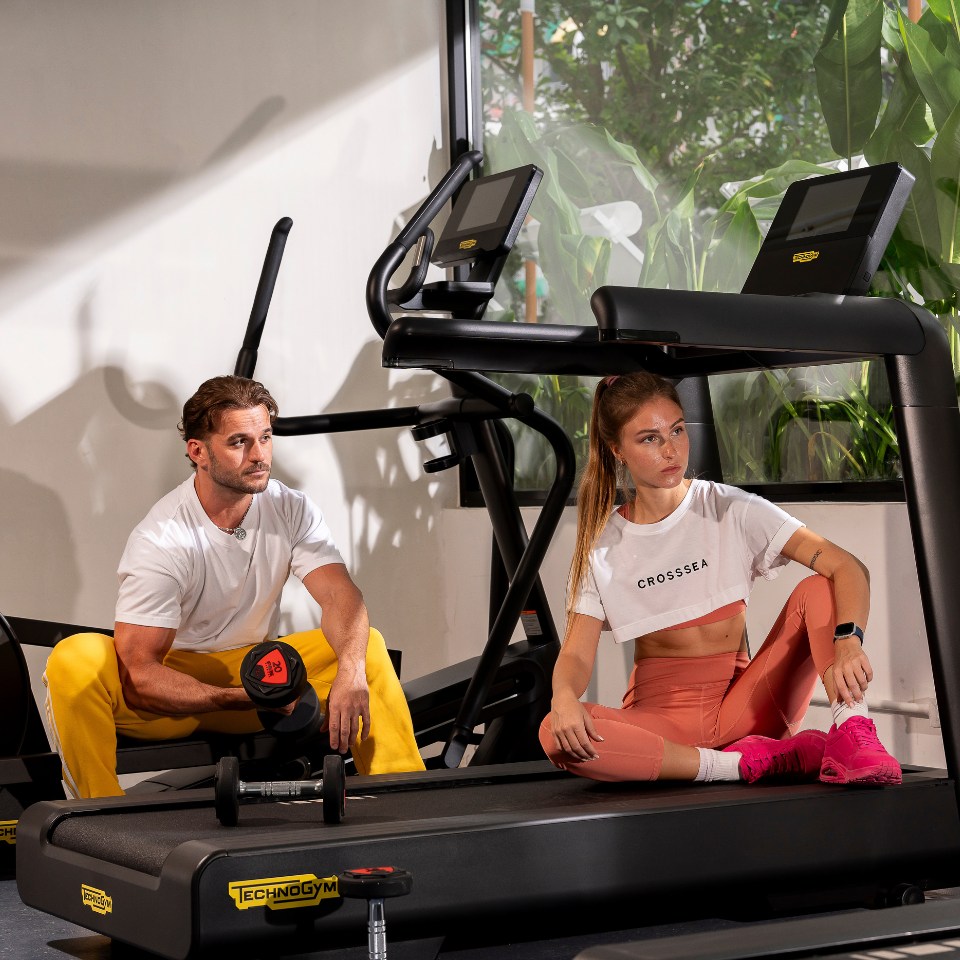 A man in a white shirt and yellow pants lifts weights, while a woman in a cropped white shirt and pink leggings sits on a treadmill. The gym setting is bright and modern.