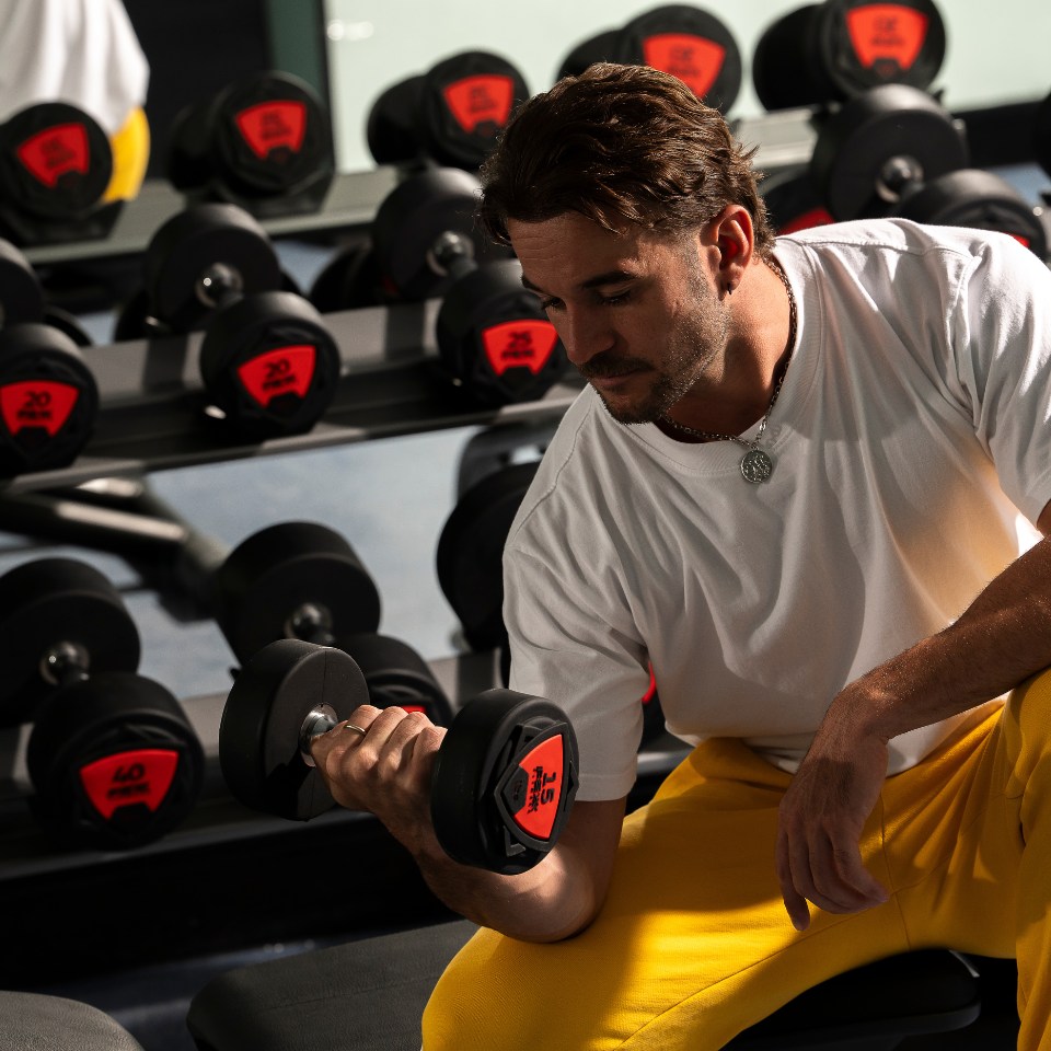 A person in a white shirt and yellow pants lifts a dumbbell in a gym. They are focused, surrounded by rows of weights, conveying determination.