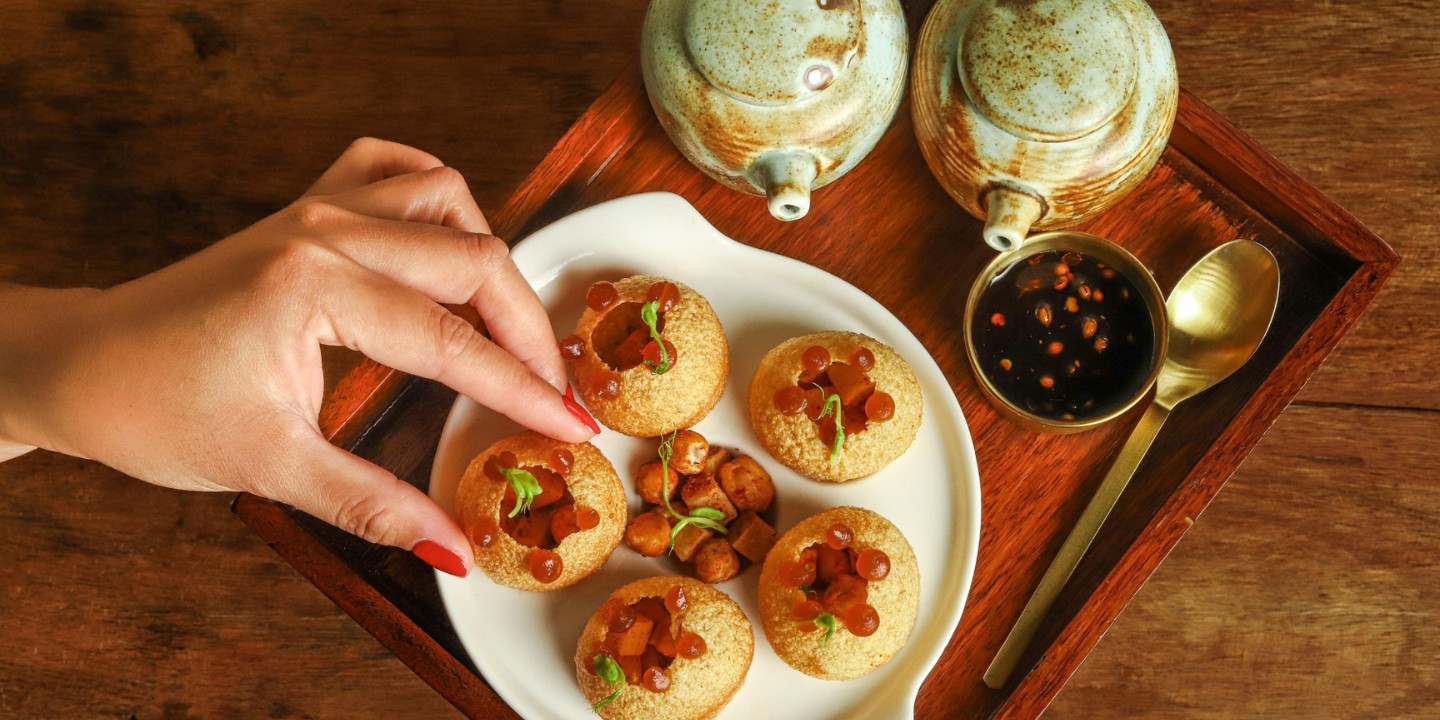 A hand with red nails reaches for a crispy, round snack on a white plate. The snacks are filled with garnishes and a side of small, diced fillings. A ceramic teapot sits nearby, creating a cozy and inviting scene.