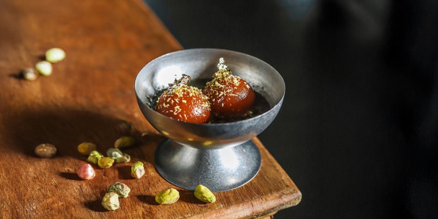 Silver bowl with two gulab jamuns, topped with gold leaf and nuts, placed on a rustic wooden table. Scattered colorful nuts surround the bowl.