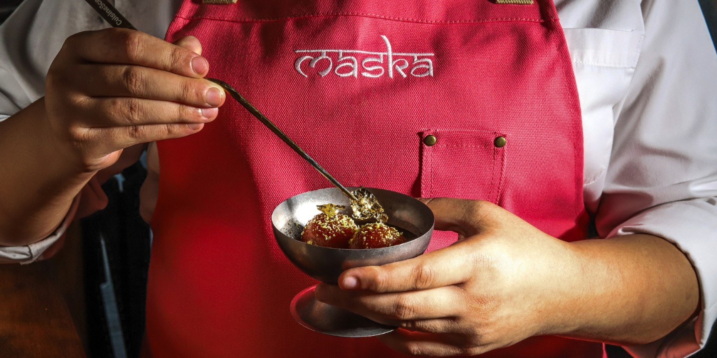 A person in a red apron holds a metal bowl with a spoon, containing dessert garnished with gold flakes. The apron has "maska" embroidered on it.