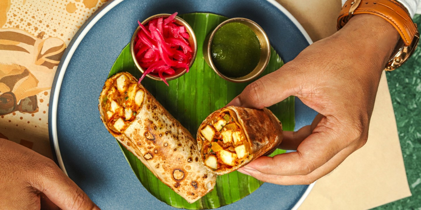 A hand holds a sliced paneer wrap on a banana leaf-lined blue plate, accompanied by bowls of green chutney and pickled onions, creating a vibrant, appetizing scene.