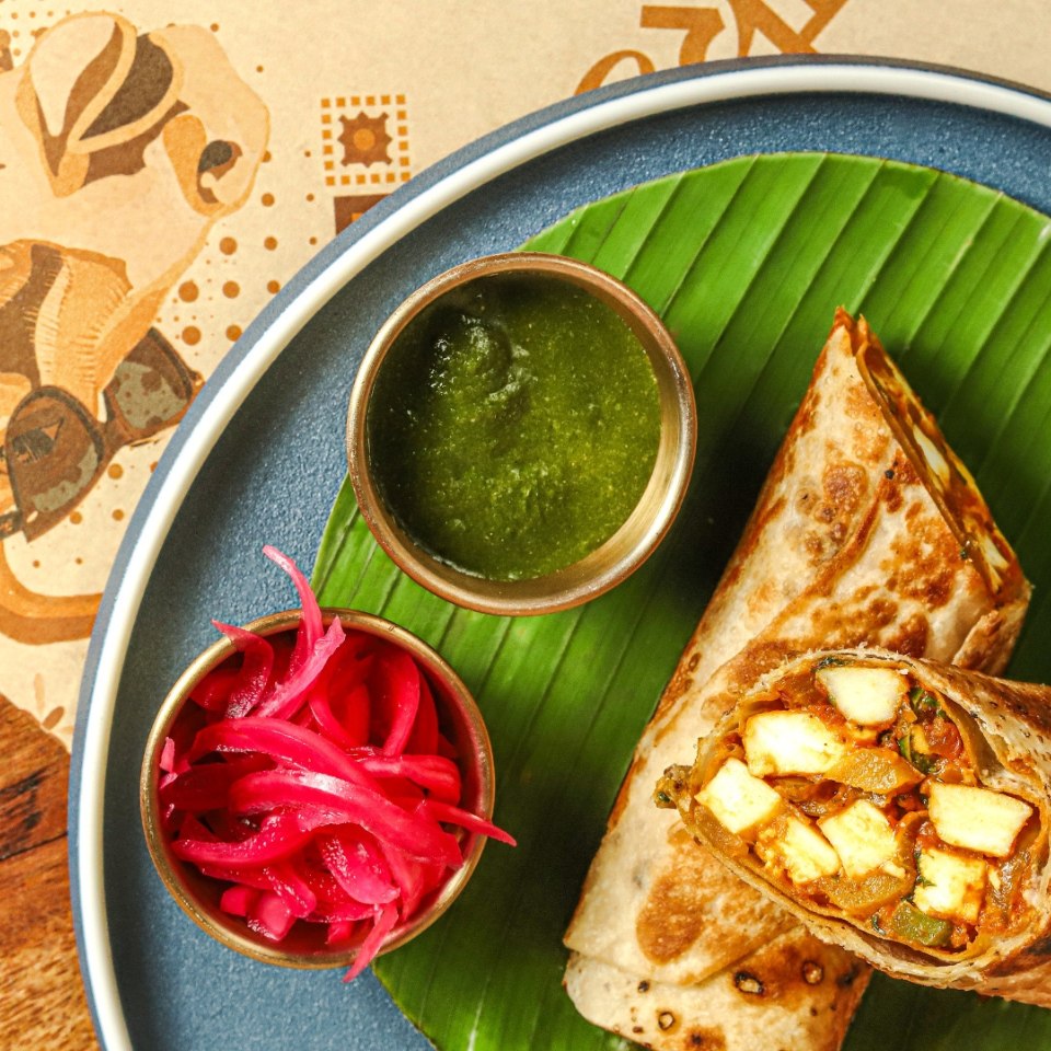 A vibrant plate featuring a paneer wrap on a banana leaf, accompanied by green chutney and pickled onions. The setting exudes a colorful, fresh vibe.
